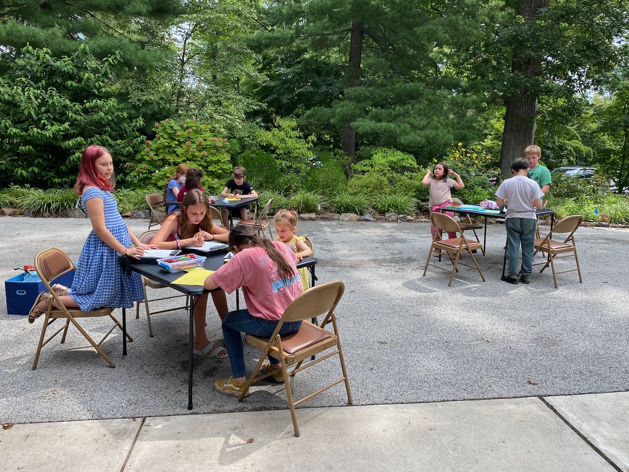 Children of a variety of ages, genders, and racial backgrounds seated around small tables outdoors participating in faith development activities