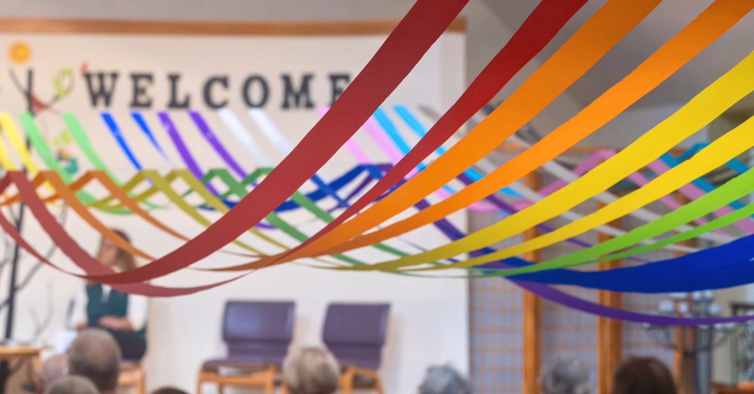 Rainbow paper streamers hang across the room at an MLUC service, with the word WELCOME on the wall behind them.
