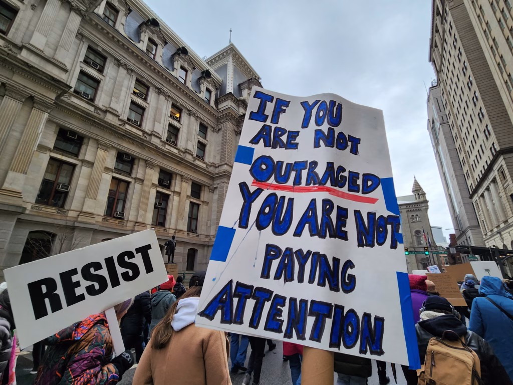 Two signs being held up in a city, one saying RESIST and a second saying If you are not outraged, you are not paying attention.