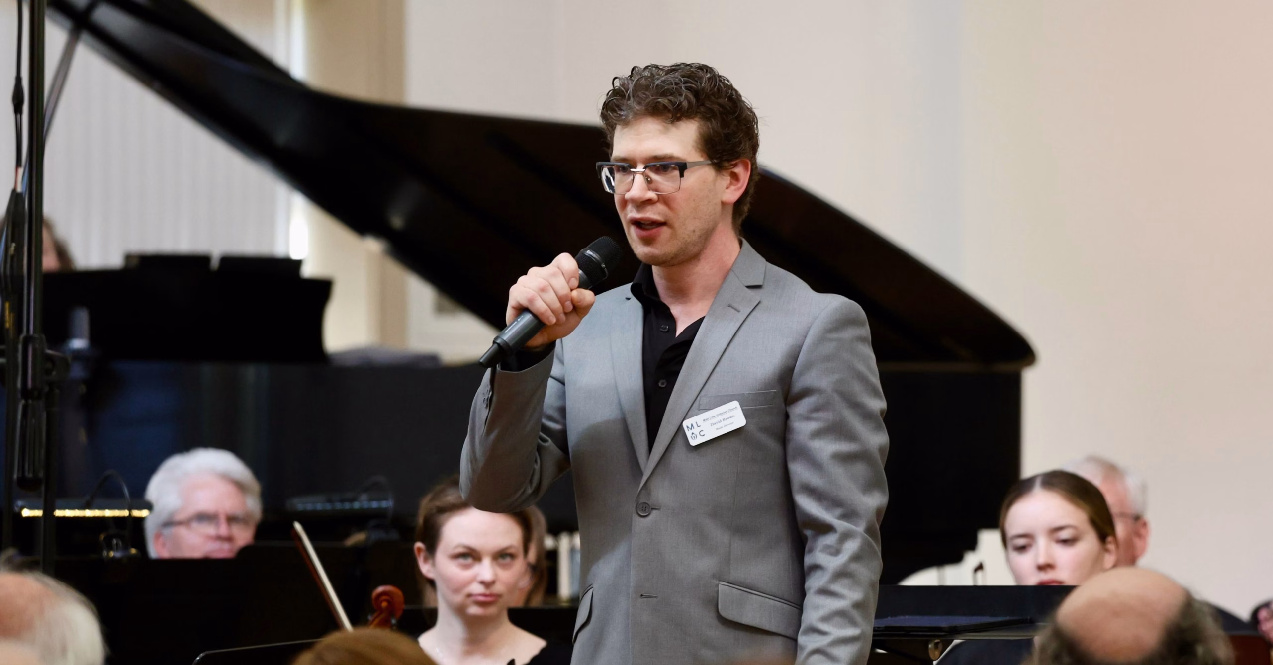 David Brown, wearing a gray suit jacket over a black shirt and glasses, holds a handheld microphone as he addresses the room. He stands in front of an open grand piano, and several seated orchestra musicians are visible in the background.