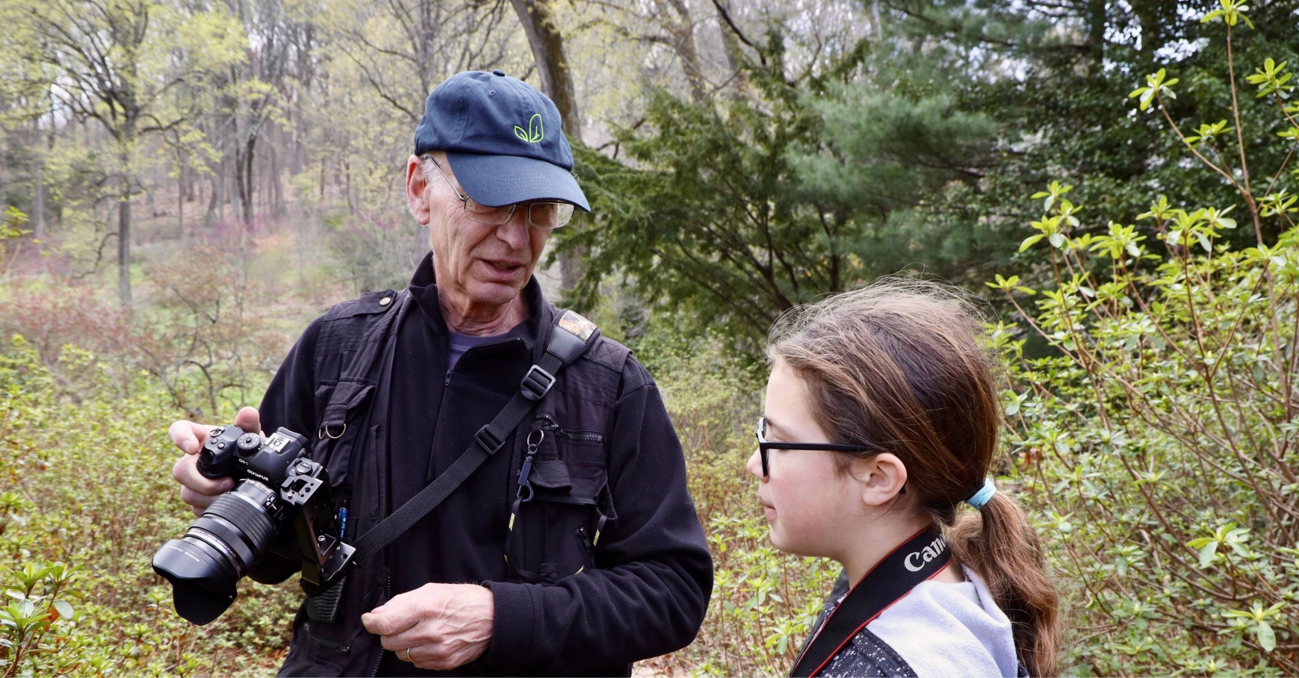 David Hucker, in a navy cap and dark fleece, shows his camera to a young student with glasses and a ponytail, who wears a camera strap around her neck, in a leafy spring garden.