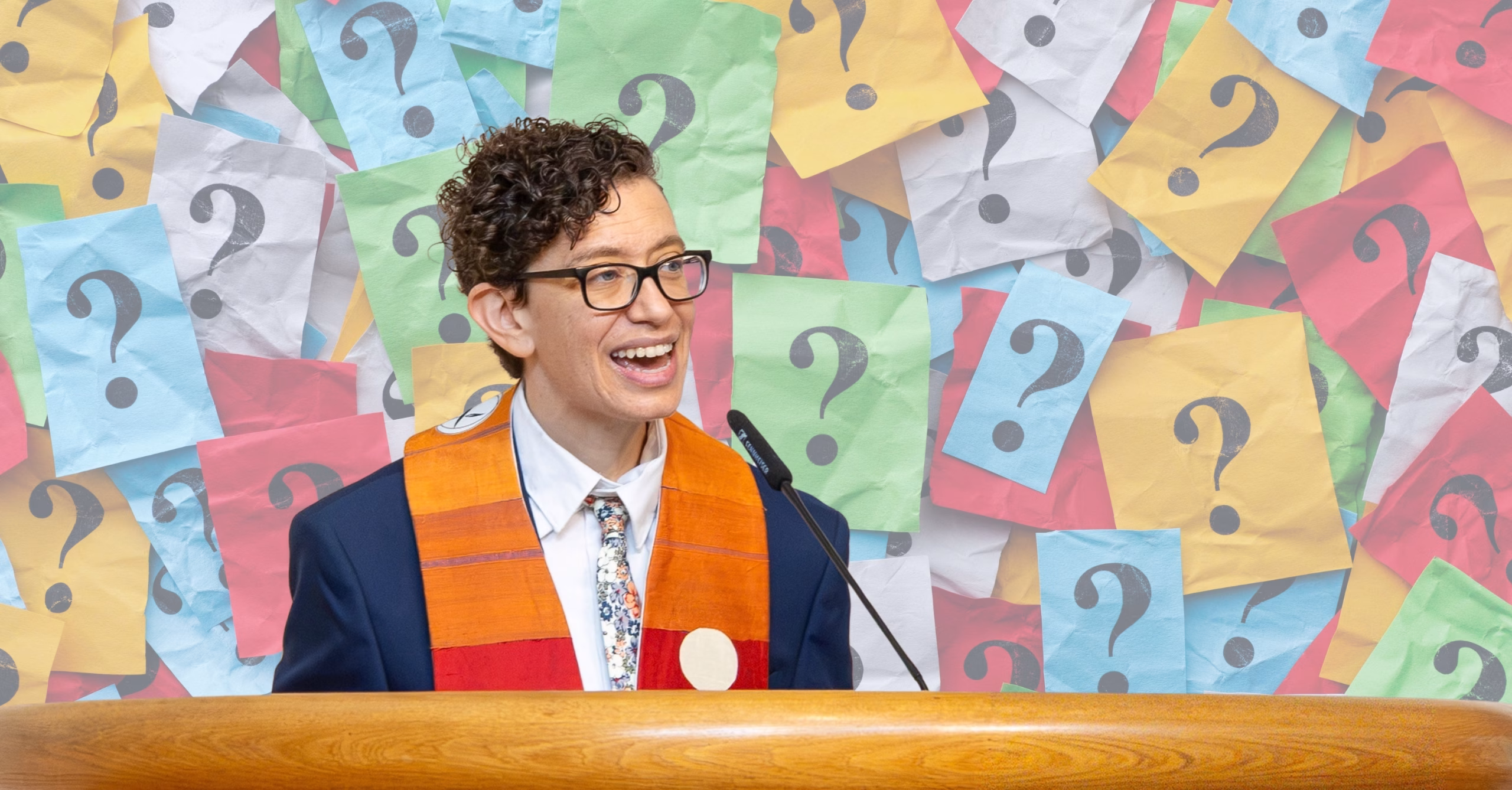 Rev. Lisa Schilansky smiles mid-speech at a wooden pulpit, wearing glasses, a navy blazer, a floral tie, and an orange-and-red stole. Behind her, a wall of colorful crumpled paper notes each bears a large question mark.